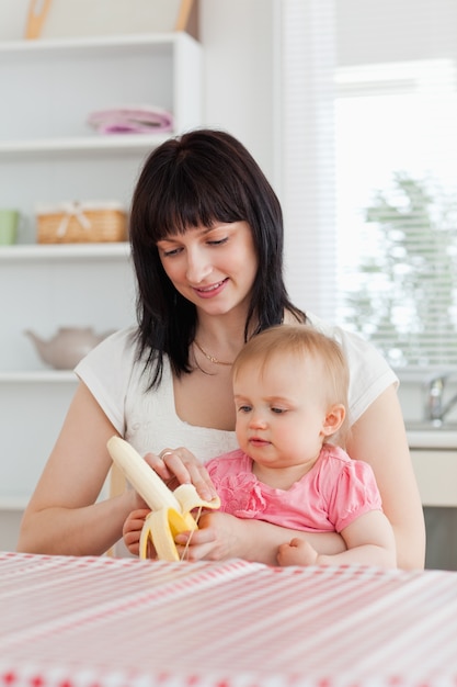 Mooie donkerbruine vrouw die een banaan pealing terwijl het houden van haar baby op haar knieën in de keuken