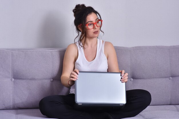 Mooie donkerbruine vrouw in rode glazen op bank die laptop met behulp van