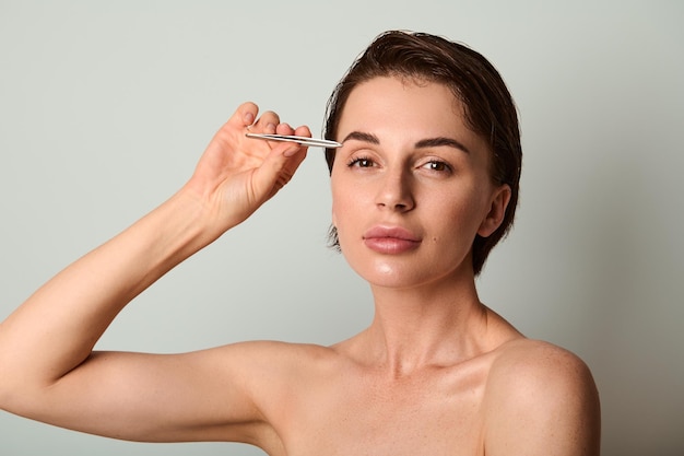 Foto mooie donkerharige blanke vrouw met stalen pincet en het verwijderen van haar uit wenkbrauwen poseren tegen lichtgrijze achtergrond met kopie advertentieruimte. schoonheidsbehandeling, verzorging, gezichts- en huidverzorging