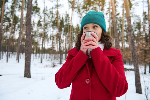 Mooie donkerharige Europese mooie vrouw in heldere rode winter wollen jas en groene gebreide muts thee of koffie drinken uit een thermomok rusten in een besneeuwd bos genieten van een weekend buiten