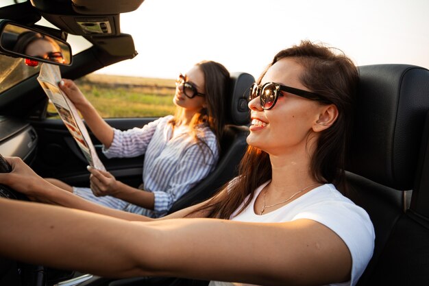 Mooie donkerharige jonge vrouwen met een zonnebril zitten op een zonnige dag in een zwarte cabriolet. Een van hen houdt de kaart in haar handen. .