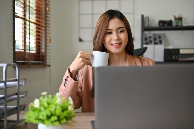 Mooie duizendjarige Aziatische zakenvrouw die naar het laptopscherm kijkt terwijl ze 's ochtends koffie drinkt