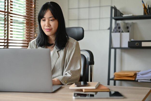 Mooie duizendjarige Aziatische zakenvrouw die zich concentreert op haar werk aan haar bureau