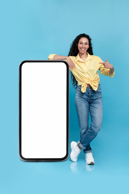 Mooie duizendjarige vrouw die mockup aanbiedt voor nieuwe mobiele app op gigantisch smartphonescherm op blauw