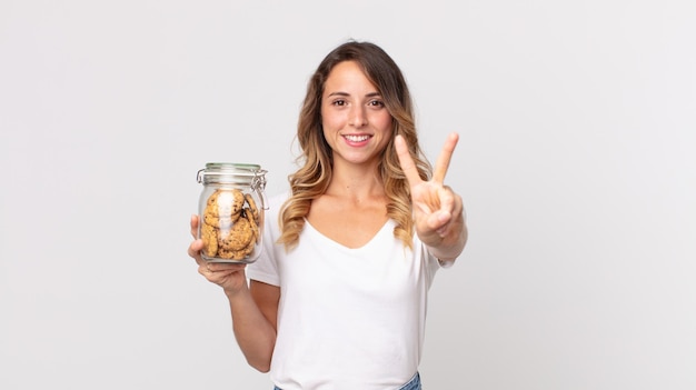 Mooie dunne vrouw die lacht en er vriendelijk uitziet, nummer twee toont en een glazen fles met koekjes vasthoudt