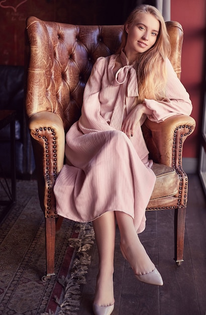 Mooie elegante blondevrouw in roze kledingszitting in leerleunstoel
