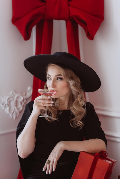 Mooie elegante vrouw in zwarte feestelijke jurk en hoed met champagneglas en nieuwjaarsgeschenken Zachte selectieve focus