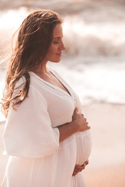 Mooie elegante zwangere vrouw die witte trendy jurk over zee draagt