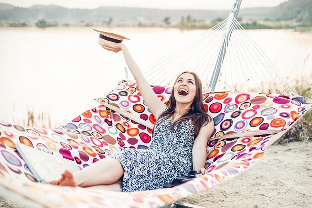 Foto mooie emotionele hipster meisje vreugde uitdrukken terwijl u ontspant in een hangmat op vakantie op het strand reis lachend meisje in boho jurk en hoed rustend in de buurt van meer