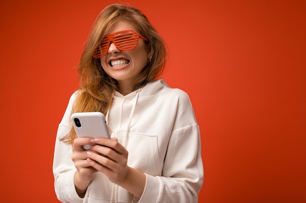 Mooie emotionele jonge blonde vrouw stijlvolle witte hoodie en grappige kleurrijke bril dragen