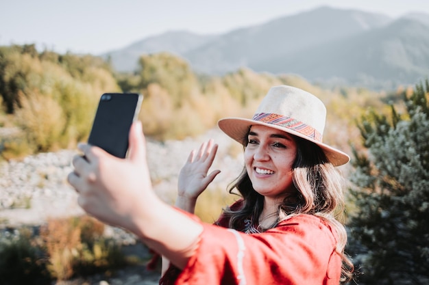 Mooie en glimlachende jonge reizigersvrouw die een videogesprek voert vanaf haar mobiel in een natuurlijke ruimte