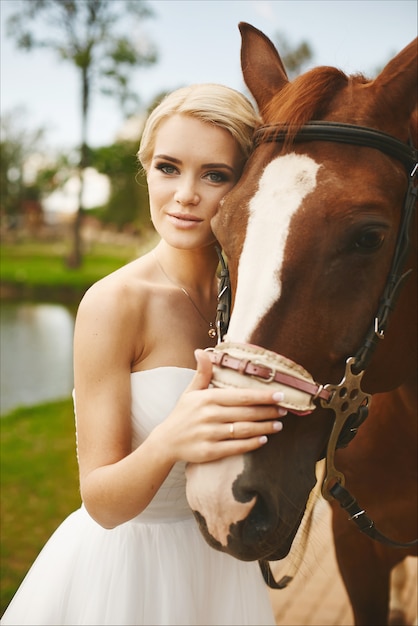 Mooie en modieuze jonge bruid, blonde model meisje met blauwe ogen en stijlvolle kapsel in witte jurk poseren met bruin paard buitenshuis