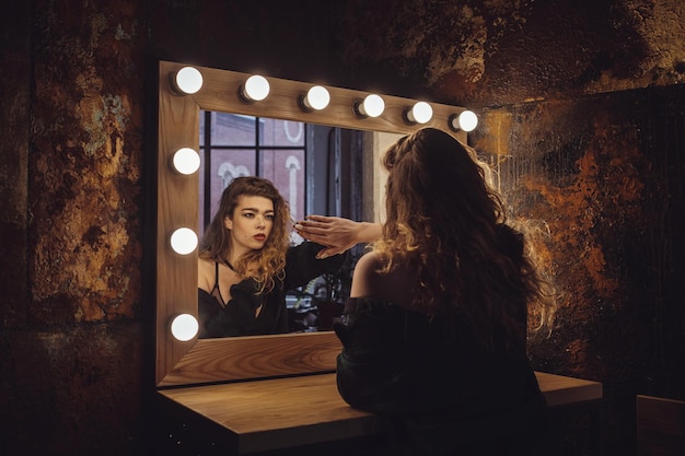 Mooie fashion model vrouw poseren in de buurt van de spiegel Vrouw in een schoonheidssalon kijkt naar haar reflectie in de spiegel met lampen en controleert kapsel en make-up