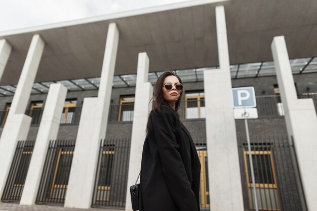 Mooie fijne jonge vrouw in trendy zonnebril in mooie zwarte jeugdblazer met stijlvolle handtas loopt in de buurt van modern gebouw in de stad. mooi mooi stedelijk meisje in mode vrijetijdskleding op straat.