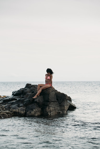 Mooie fit zwarte vrouw zittend in rotsen in bikini aan de kust