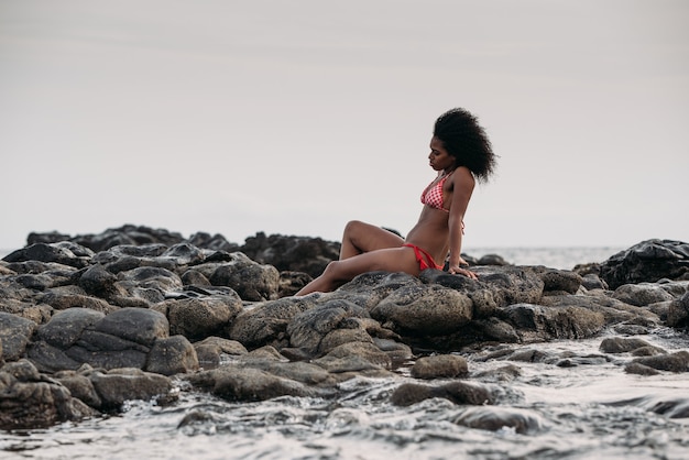 Mooie fit zwarte vrouw zittend in rotsen in bikini aan de kust