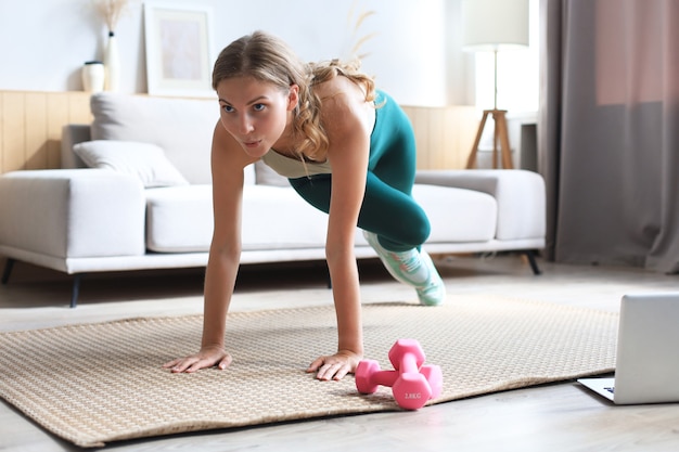 Mooie fitness vrouw doet bergbeklimmer oefeningen kijken naar online tutorials op laptop, training in de woonkamer. Gezonde levensstijl. Meisje gaat thuis sporten.