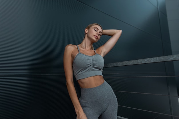 Mooie fitness vrouw model met mooi lichaam in trendy grijze sportkleding met tank top bh en legging staat en geniet in de buurt van moderne donkere metalen wand