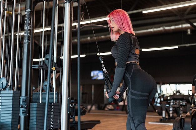 Mooie fitnessvrouw met roze haar in een zwart trainingspak traint en pompt spieren op de simulator in de sportschool