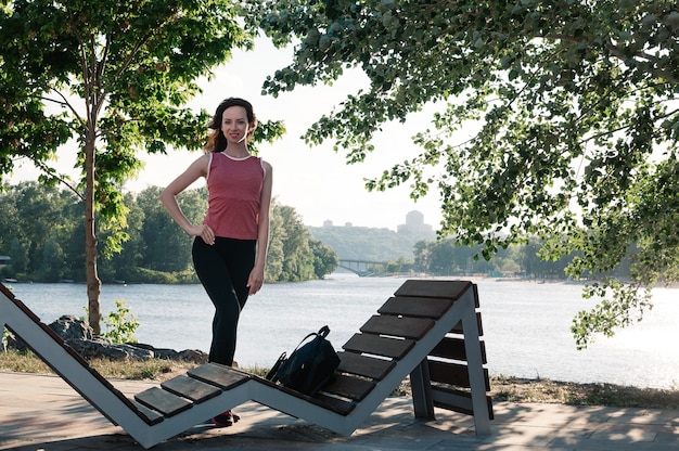 Foto mooie fitte jonge vrouw die bij de bank staat en strakke trainingskleding draagt die buiten glimlacht