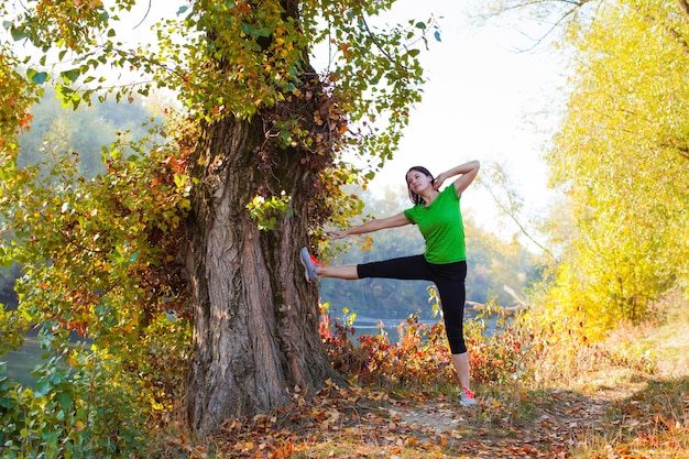 Mooie fitte vrouw die haar benen en armen strekt en zich voorbereidt op de ochtendrun in het herfstbos met kleurrijke bomen en rivier.