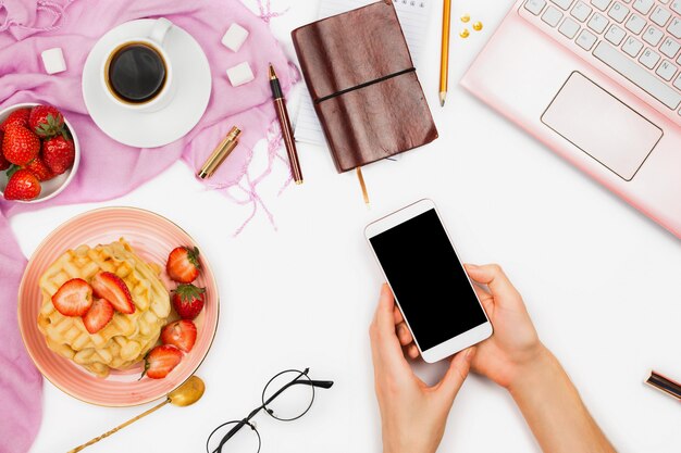 Mooie flatlay-regeling met kop koffie, hete wafels met room en aardbeien, laptop en de holdingssmartphone van de vrouwenhand: concept bezig ochtendontbijt, witte achtergrond.