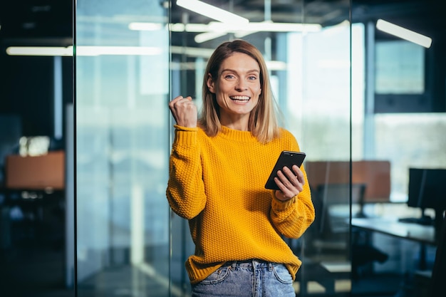 Mooie freelancer-vrouw kijkt naar de camera en glimlacht, verheugt zich over het winnen en winnen online met een telefoon en het vieren van triomf