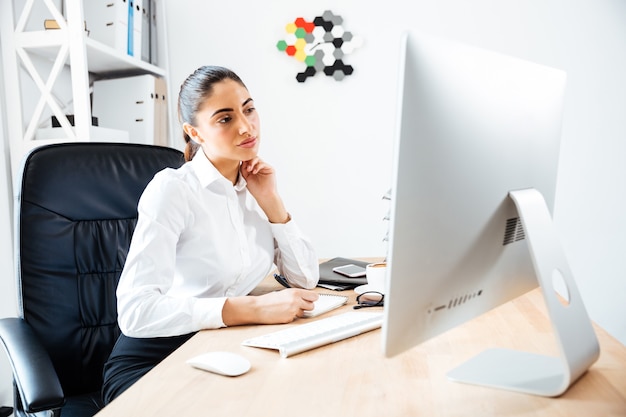 Mooie geconcentreerde zakenvrouw die aantekeningen maakt en naar het computerscherm kijkt terwijl ze op kantoor zit