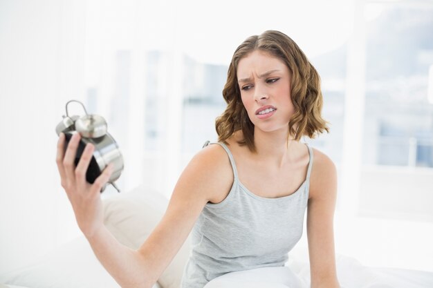 Mooie geërgerde vrouw die een wekker houdt die het bekijkt zittend op haar bed