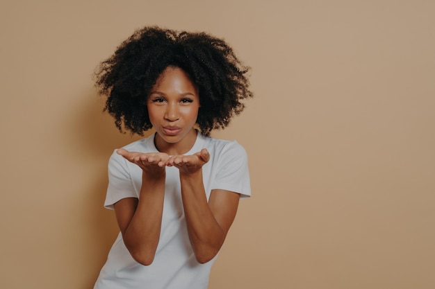 Mooie gekrulde flirterige afrikaanse vrouw die luchtkus verzendt en glimlacht terwijl ze poseert op beige achtergrond