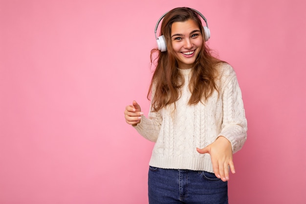 Mooie gelukkig lachende jonge brunette krullend vrouw dragen witte trui geïsoleerd op roze achtergrond muur dragen witte bluetooth koptelefoon luisteren naar coole muziek kijken naar camera