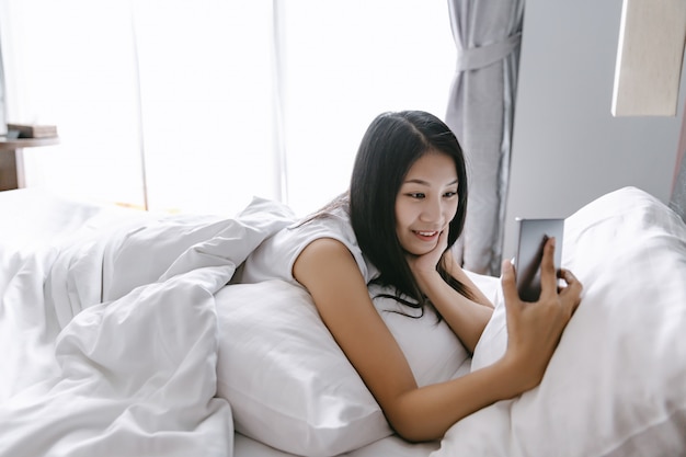 Mooie gelukkige Aziatische vrouw met behulp van mobiele telefoon voor videogesprek op bed na wakker worden in moderne slaapkamer thuis, sociale afstand nemen en nieuw normaal concept