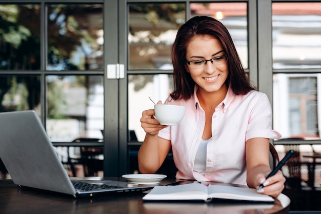 Mooie, gelukkige brunette houdt een kop in haar hand, werkt op een laptop in een café.