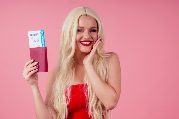 Mooie gelukkige jonge blonde vrouw die een zomerjurk draagt die over een roze achtergrond in de studio staat en een paspoort met vliegtickets toont.