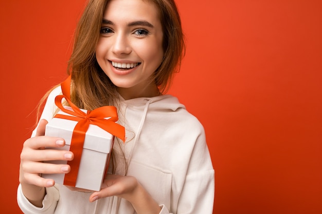 Mooie gelukkige jonge blonde vrouw die over kleurrijke muur wordt geïsoleerd die modieuze vrijetijdskleding draagt die giftdoos houdt en camera bekijkt