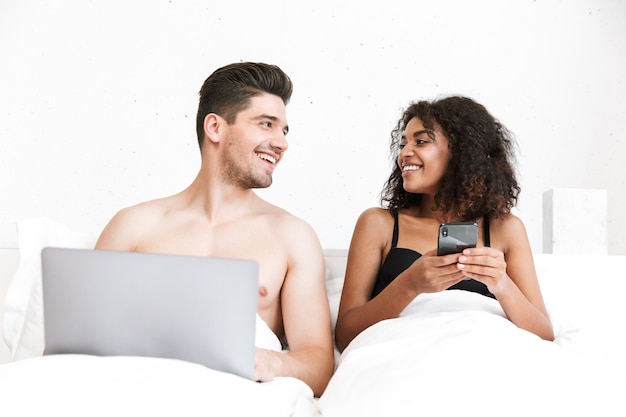 Mooie gelukkige jonge multi-etnisch paar ontspannen in bed onder deken, man met laptopcomputer, vrouw met mobiele telefoon