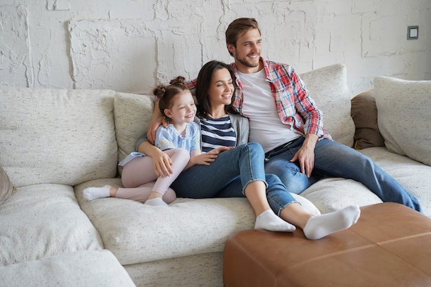 Mooie gelukkige jonge ouders en hun dochtertje tv kijken en glimlachen terwijl ze thuis op de bank zitten.