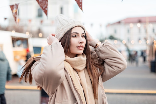 Mooie gelukkige jonge stijlvolle vrouw in modieuze winter bovenkleding in een vintage gebreide muts met een jas en sjaal loopt op de beurs. wintervakantie buiten