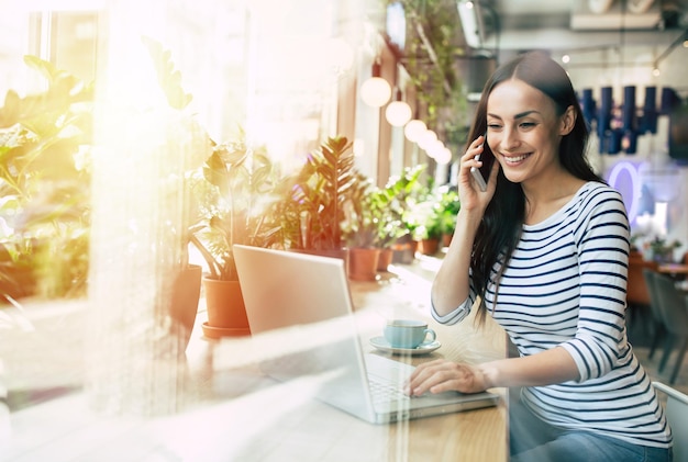 Mooie gelukkige jonge vrouw die laptop en smartphone gebruikt terwijl ze tijdens de lunch een kopje koffie drinkt in café