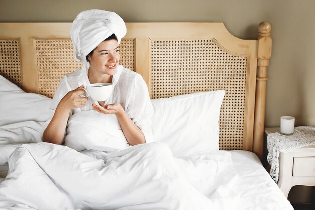 Mooie gelukkige jonge vrouw koffie drinken in bed in hotelkamer of thuis slaapkamer Stijlvolle brunette meisje in witte handdoek genieten van ochtend met warme drank Ruimte voor tekst