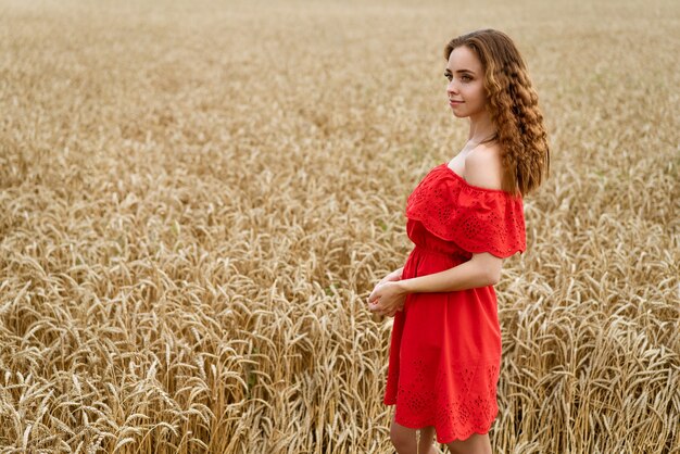 Mooie gelukkige jonge vrouw met krullend haar in een rode jurk poseren in een tarweveld