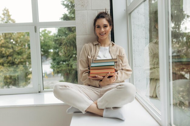 Mooie gelukkige jonge vrouw zittend op de vensterbank met een boek in haar handen