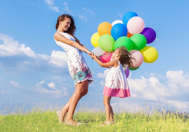 Mooie gelukkige moeder met dochter die plezier heeft in het groene veld met kleurrijke ballonnen