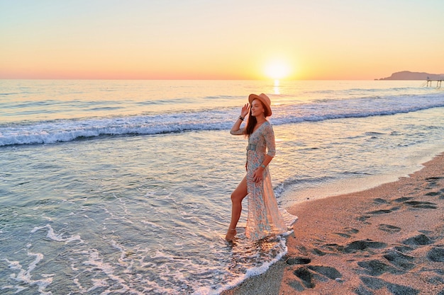 Mooie gelukkige romantische schattige boho-meisjesreiziger die een lange jurk en hoed draagt die alleen staat aan de kust met schuimgolven zee bij zonsondergang