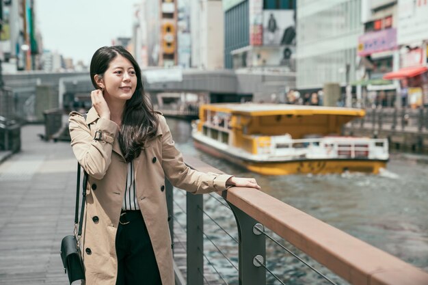 Mooie gelukkige vrouw die in de buurt van de osaka-rivier loopt door dotonbori en shinsaibashi en opzij kijkt. lachende dame veegt haar en houdt de leuning vast in het drukke stadsgebied. vrolijke vrouw geniet van zonlicht.