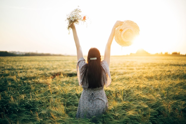 Mooie gelukkige vrouw met wilde bloemen die geniet van de zonsondergang in een gerstveld Atmosfeer rustig moment langzaam leven Stijlvol vrouwelijk bloemen verzamelen en ontspannen in de avond zomer platteland