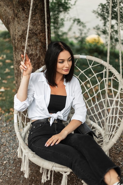 Mooie gelukkige zakenmodelvrouw in modekleding met een wit overhemd en zwarte spijkerbroek zit en ontspant op een gebreide schommel in de tuin