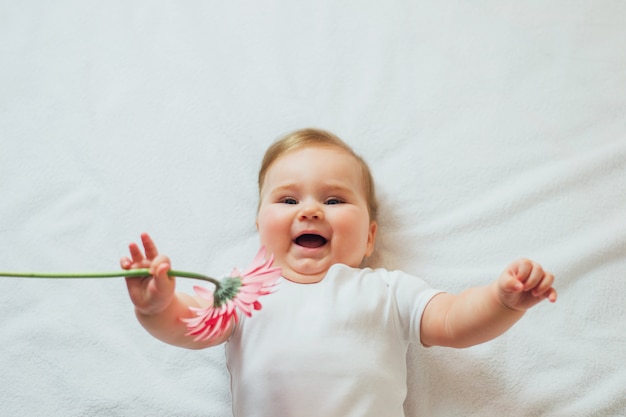 Mooie gelukkige zuigelingsbaby die op witte bladen ligt die een bloem houden. Gelukkig kind met bloem die witte bodysuit draagt.