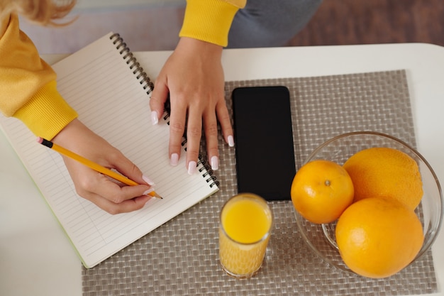 Mooie gemanicuurde handen van jonge vrouw schrijven in planner op salontafel met smartphone en glas vers sinaasappelsap