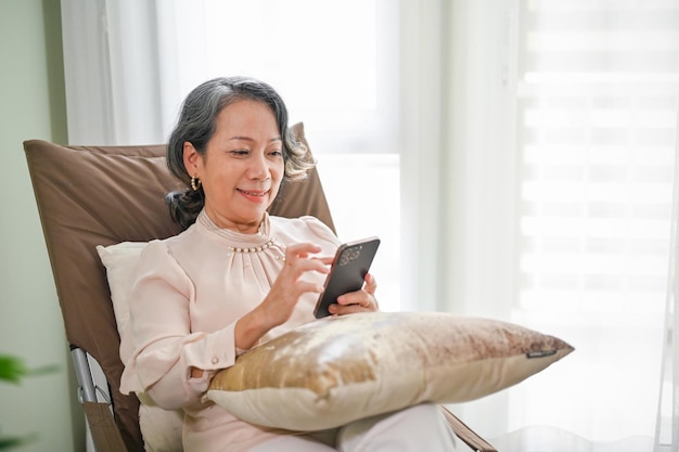 Mooie gepensioneerde aziatische vrouw uit de jaren 60 die haar smartphone gebruikt terwijl ze ontspant in haar comfortabele fauteuil